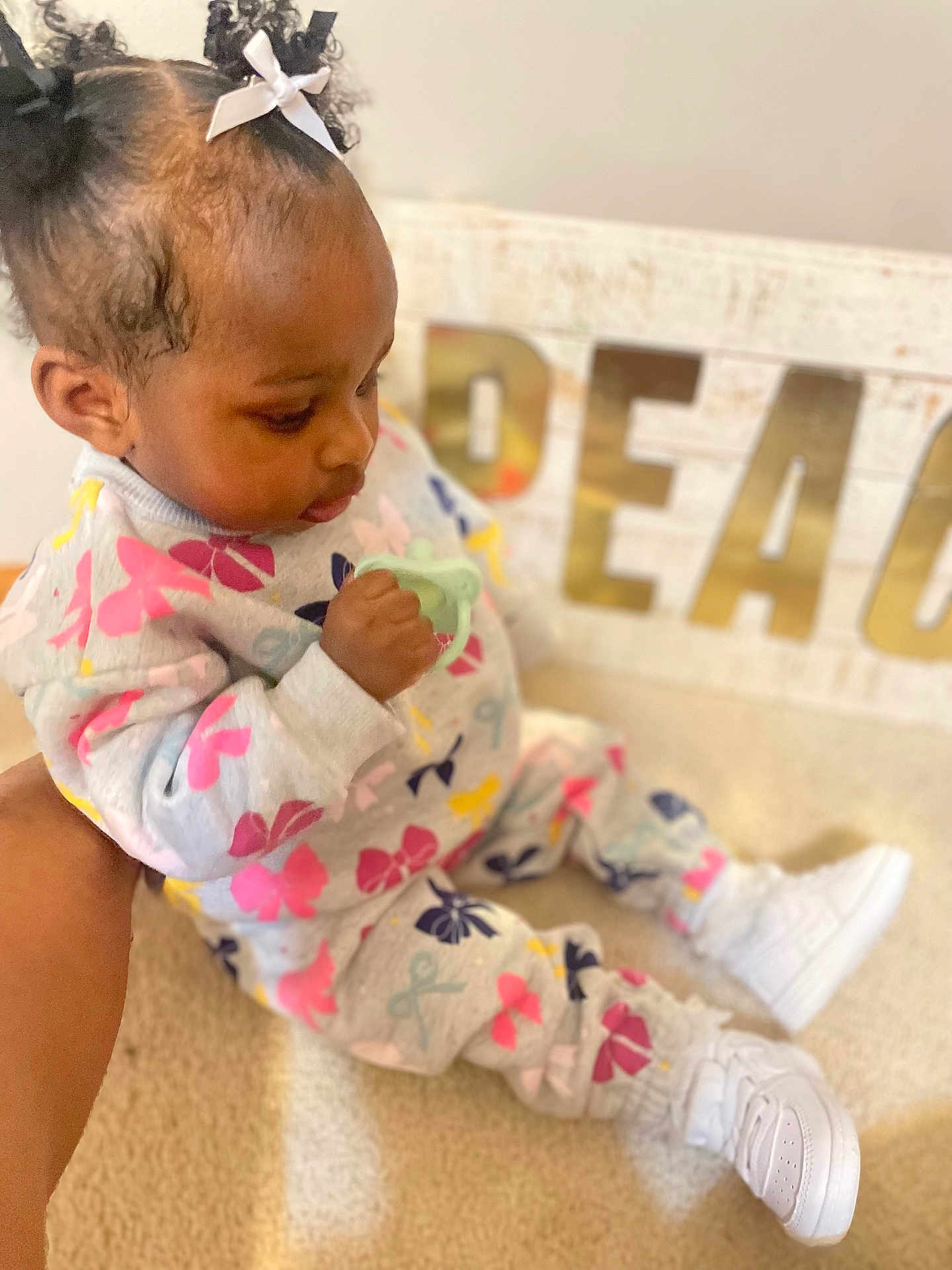 Aunystee Star is registered to the contest to win money with this photo: toddler, child, indoor, carpet, pacifier, curly_hair, bows, sneakers, colorful_clothing, sitting, person, young_child, baby, cute, holding_object, floor, soft_lighting, blurred_background, peace_sign, portrait