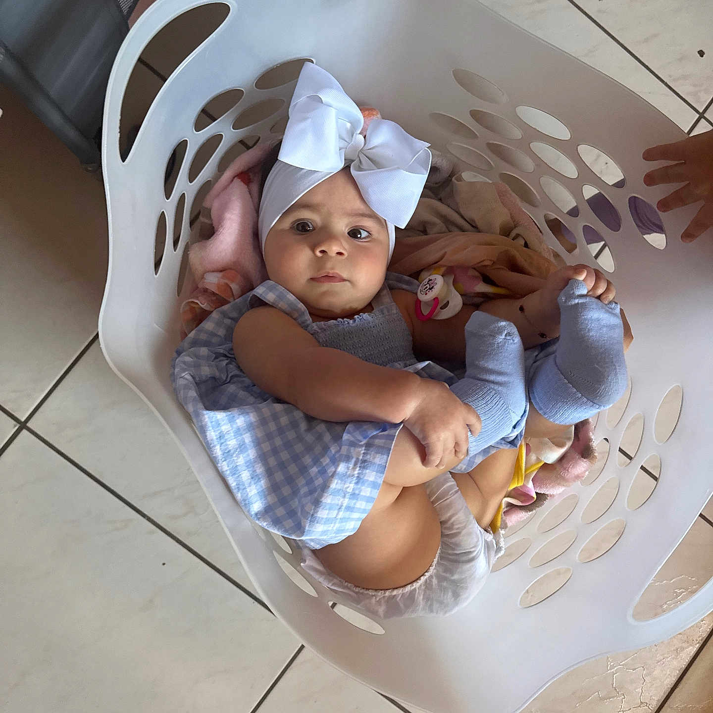 Alyssa is registered to the contest to win money with this photo: baby, child, laundry_basket, white_bow, blue_dress, socks, blanket, tile_floor, indoors, pacifier, hand, cute, infant, clothing, person, looking_at_camera, basket, soft_toys, floor, playful