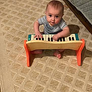Titus is registered to the contest to win money with this photo: baby, infant, toy_piano, musical_instrument, carpet, patterned_rug, bed, quilt, bedframe, sitting, barefoot, onesie, striped_clothing, face, eyes, hands, playing, indoor, cute, looking_up