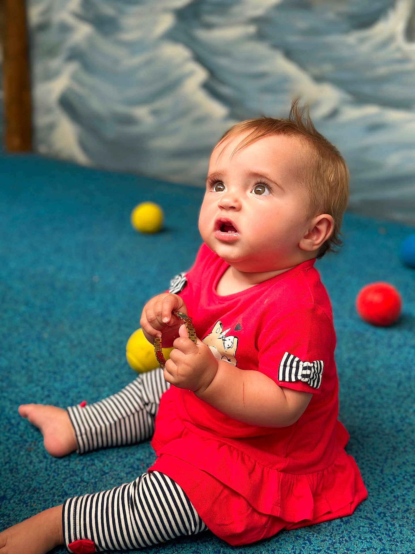 Giulia a rejoint le concours — aidez-le/la à gagner de superbes lots ! baby, child, sitting, red_dress, striped_leggings, bracelet, blue_carpet, yellow_ball, orange_ball, blue_ball, curious_expression, indoor, floor, person, cute, young_child, play, toy, portrait, casual_clothing