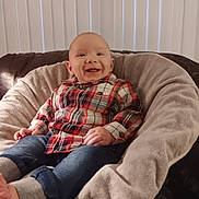 Saint is registered to the contest to win money with this photo: baby, child, smiling, plaid_shirt, jeans, cozy, chair, cushion, indoor, portrait, happy, infant, face, clothing, comfortable, casual, cute, young, relaxed, home