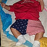 Elijah is registered to the contest to win money with this photo: arms_raised, baby, bed, bedroom, blanket, bottle, child, clothing, face, flag_shorts, indoor, infant, lying_down, patriotic, portrait, quilt, red_shirt, remote_control, sleeping, socks