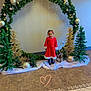 child, girl, holiday, christmas, red_dress, decorations, arch, greenery, ornaments, fairy_lights, reindeer, small_trees, indoor, floor_tiles, festive, smiling, holiday_season, cute, heart_doodle, celebration