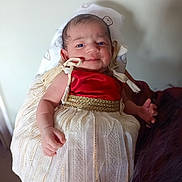 Jennifer is registered to the contest to win money with this photo: baby, child, infant, dress, red_top, white_skirt, golden_belt, smile, hand, face, person, cute, indoors, soft_background, holding, fabric, portrait, newborn, human, skin