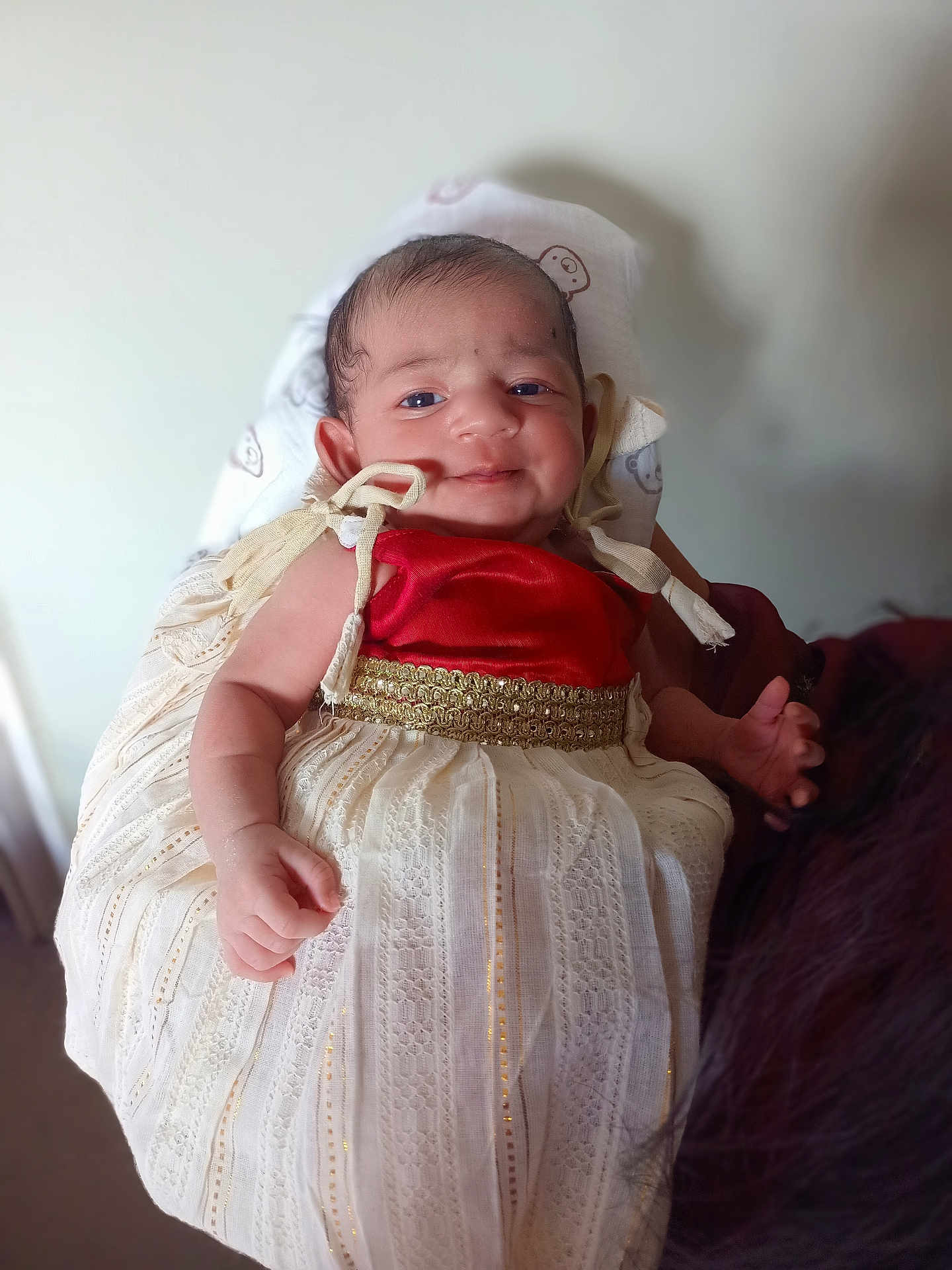 Jennifer is registered to the contest to win money with this photo: baby, child, infant, dress, red_top, white_skirt, golden_belt, smile, hand, face, person, cute, indoors, soft_background, holding, fabric, portrait, newborn, human, skin