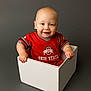 Briggs is registered to the contest to win money with this photo: baby, infant, child, smile, sitting, white_box, red_shirt, ohio_state, logo, portrait, studio, gray_background, cute, bald, hands, eyes, face, young_child, box, clothing