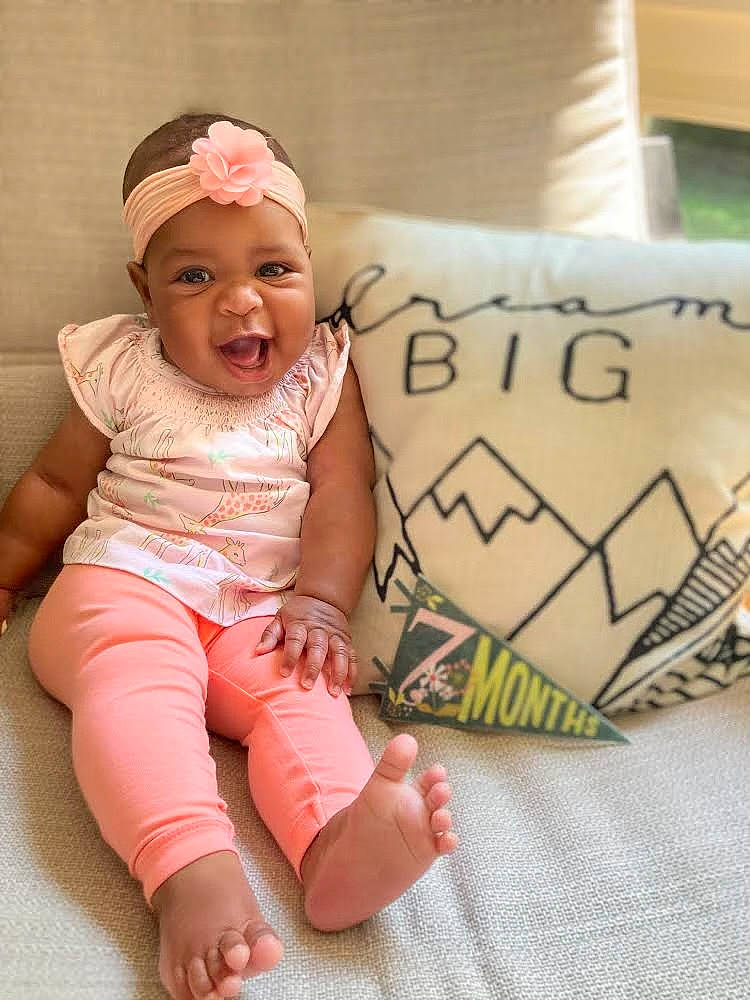 Mercedes is registered to the contest to win money with this photo: baby, baby_toddler_clothing, cheek, comfort, eye, facial_expression, flooring, happy, headwear, human, linens, mammal, person, pink, product, skin, sleeve, smile, t_shirt, textile