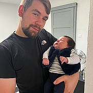 Ana participe au concours pour gagner de l'argent avec cette photo : man, baby, holding, indoor, casual, black_tshirt, sleeping_baby, beard, short_hair, striped_shirt, vest, peaceful, home_interior, door, fan, hand, person, portrait, parent, infant