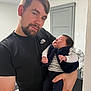 man, baby, holding, indoor, casual, black_tshirt, sleeping_baby, beard, short_hair, striped_shirt, vest, peaceful, home_interior, door, fan, hand, person, portrait, parent, infant