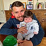 man, baby, table, wooden_furniture, bottle, family_photos, rose, portrait, indoor, smile, beard, clothing, child, person, face, hand, cabinet, cozy, home, casual