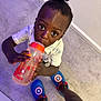 child, toddler, sippy_cup, carpet, indoor, topknot, blue_slippers, captain_america, shorts, curious, looking_up, holding, drinking, person, floor, casual_clothing, playful, wide_eyes, young_child, home