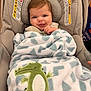 baby, infant, car_seat, blanket, smiling, tongue_out, headrest, warning_label, plush_crocodile, patterned_blanket, cozy, portrait, indoor, retail_store, hand, hair, blue_eyes, cheeky_expression, soft_fabric, adorable