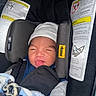 baby, car_seat, infant, blanket, hat, face, child, clothing, safety_label, seatbelt, smiling, tongue_out, cute, indoors, person, newborn, soft_texture, blue, black, white