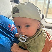 Loann participe au concours pour gagner de l'argent avec cette photo : baby, pacifier, cap, indoor, person, hand, clothing, blue_shirt, green_shirt, face, eyes, infant, holding, couch, window, curtain, adult_hand, closeup, portrait, soft_lighting