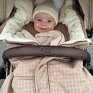 Theo is registered to the contest to win money with this photo: baby, stroller, hat, knit_hat, pom_poms, blanket, smiling, child, warm_clothing, cozy, infant, fur_lining, outdoor, person, face, cute, happy, wrapped, seat, soft