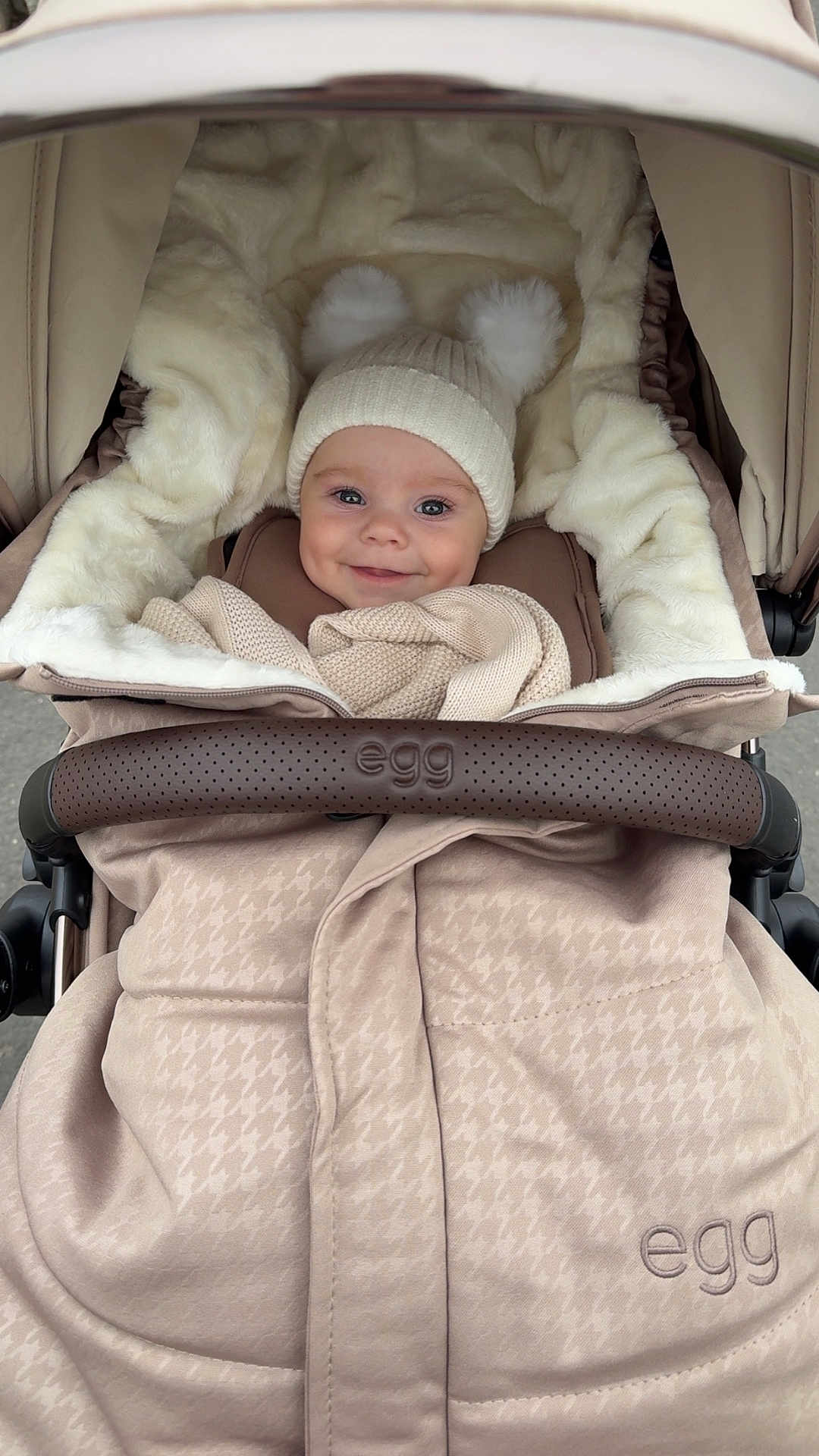 Theo is registered to the contest to win money with this photo: baby, stroller, hat, knit_hat, pom_poms, blanket, smiling, child, warm_clothing, cozy, infant, fur_lining, outdoor, person, face, cute, happy, wrapped, seat, soft