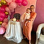 baby, woman, birthday_cake, high_chair, balloons, party, celebration, dress, smile, indoor, flooring, decorations, pink, gold, white, fabric, crown, person, glasses, happy