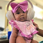 Mi Lani is registered to the contest to win money with this photo: baby, headphones, pink_bow, bib, onesie, infant, chubby, sitting, indoor, cute, child, person, apparel, clothing, headwear, accessory, soft_lighting, portrait, baby_girl, closeup