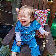 Inaya participe au concours pour gagner de l'argent avec cette photo : toddler, child, barefoot, blue_clothing, wooden_chair, colorful_pillow, outdoor, garden, plants, laughing, happy, smiling, hair, face, sitting, wood, natural_light, greenery, fence, casual