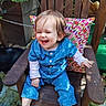 toddler, child, barefoot, blue_clothing, wooden_chair, colorful_pillow, outdoor, garden, plants, laughing, happy, smiling, hair, face, sitting, wood, natural_light, greenery, fence, casual