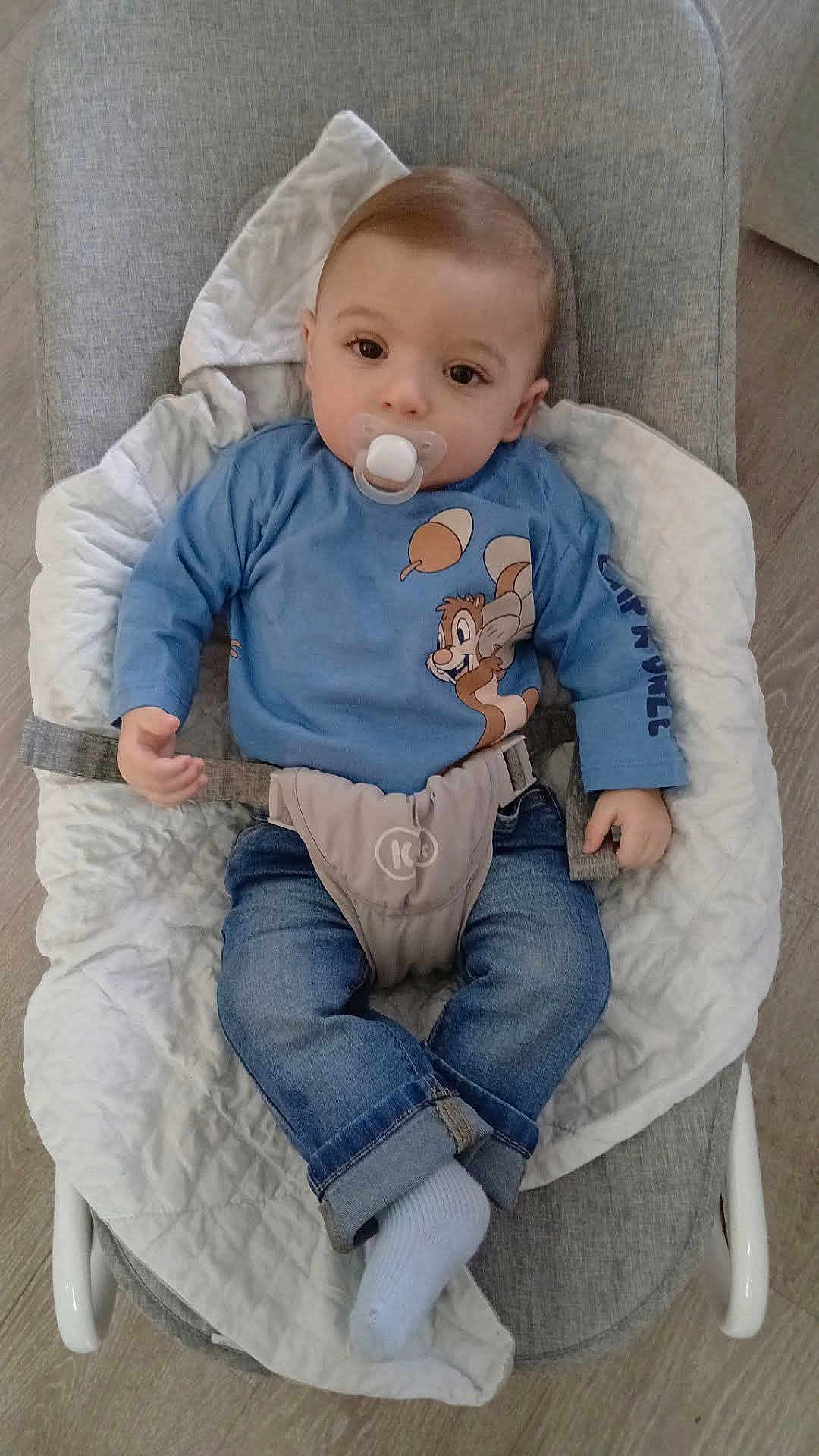 Zarek a rejoint le concours — aidez-le/la à gagner de superbes lots ! baby, blanket, bouncer, brown_hair, cartoon_print, cute, face, hand, indoor, infant, jeans, looking_at_camera, onesie, pacifier, portrait, safety_belt, sitting, sock, strap, toddler