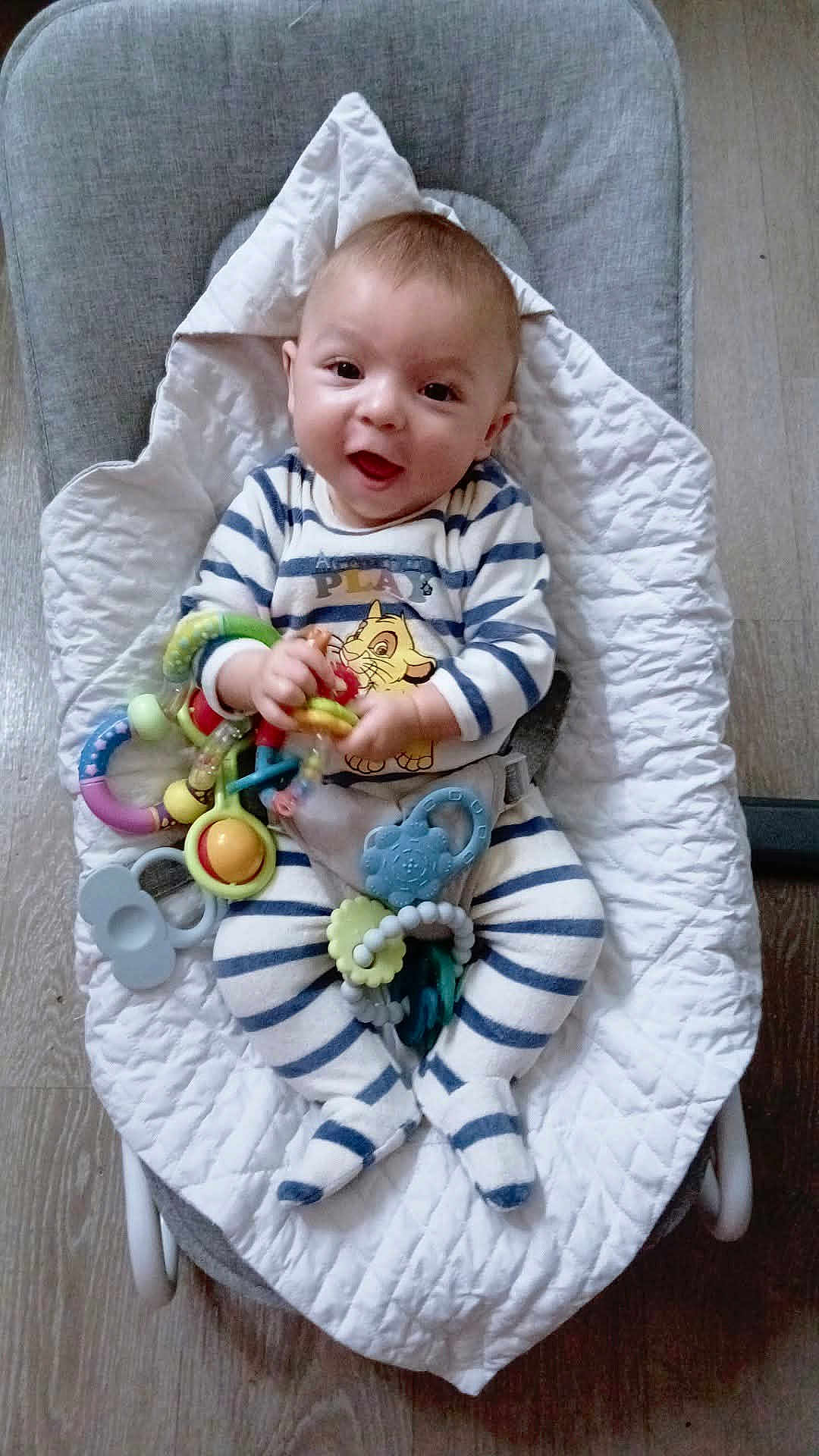 Zarek a rejoint le concours — aidez-le/la à gagner de superbes lots ! baby, baby_seat, child, clothing, colorful, comfort, cushion, cute, floor, happy, holding, indoor, infant, lying_down, person, playful, small, smiling, striped_pajamas, toy