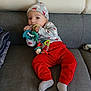 baby, child, cute, face, festive_clothing, gray_couch, gray_hat, headwear, home, indoor, infant, person, playing, red_pants, relaxed, seated, smiling, socks, teething_toy, toy