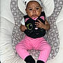 baby, infant, child, headband, bow, pink_pants, black_bib, cat_design, socks, patterned_seat, cushion, indoor, person, cute, portrait, seated, small_hands, looking_at_camera, clothing, soft_fabric