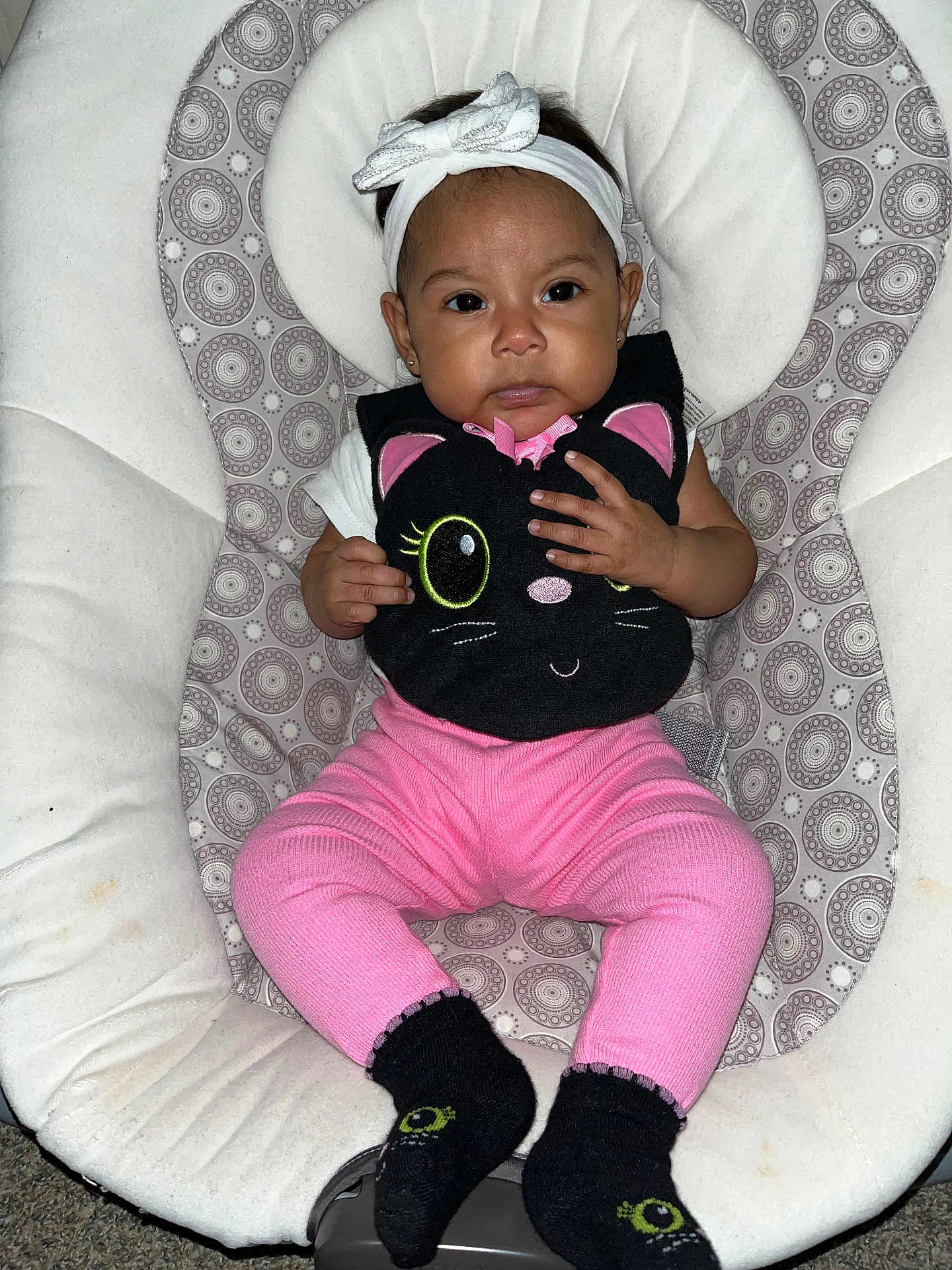 Maya joined the competition — help win amazing prizes! baby, infant, child, headband, bow, pink_pants, black_bib, cat_design, socks, patterned_seat, cushion, indoor, person, cute, portrait, seated, small_hands, looking_at_camera, clothing, soft_fabric
