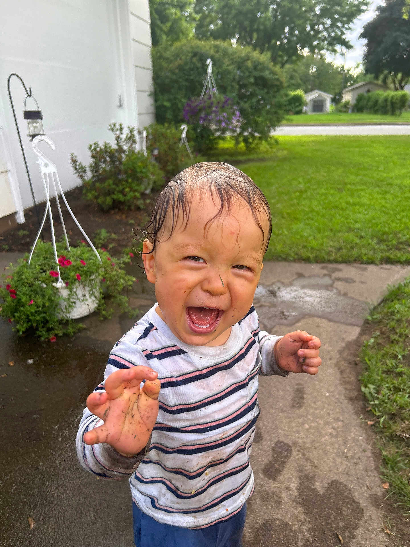 Gabriel is registered to the contest to win money with this photo: toddler, child, mud, wet_hair, striped_shirt, laughing, outdoor, sidewalk, greenery, flower_pot, grass, happy, dirt, playful, person, smiling, wet_clothes, nature, daylight, fun