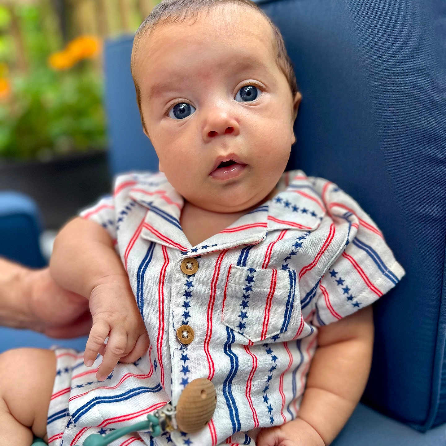 Yariel is registered to the contest to win money with this photo: baby, blue_cushion, blurred_background, child, clothing, cute, expression, face, garden, hand, infant, outdoor, pacifier_clip, portrait, relaxed, seated, stars_pattern, striped_clothing, wooden_button, young