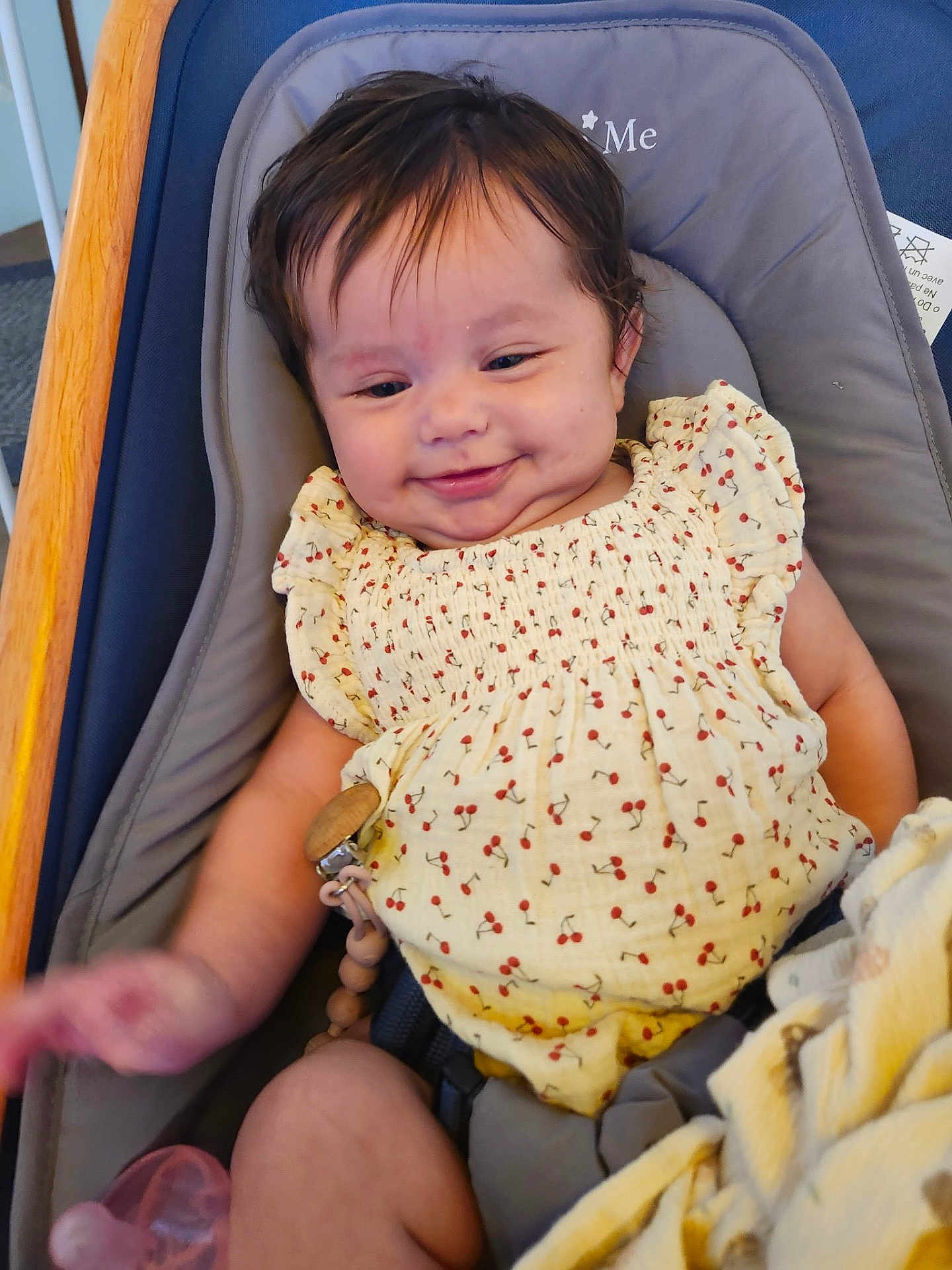 Emily joined the competition — help win amazing prizes! baby, baby_seat, blanket, cherry_pattern, child, comfort, cushion, cute, dark_hair, face, happy, indoor, infant, pacifier, person, relaxed, small_hands, smiling, wooden_frame, yellow_clothing