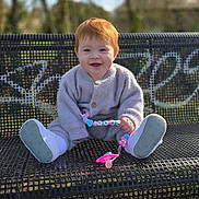 Théona participe au concours pour gagner de l'argent avec cette photo : baby, toddler, red_hair, smiling, bench, park, outdoors, grass, trees, sweater, shoes, pacifier, toy, pacifier_clip, sitting, portrait, bokeh, sunlight, cute, happy
