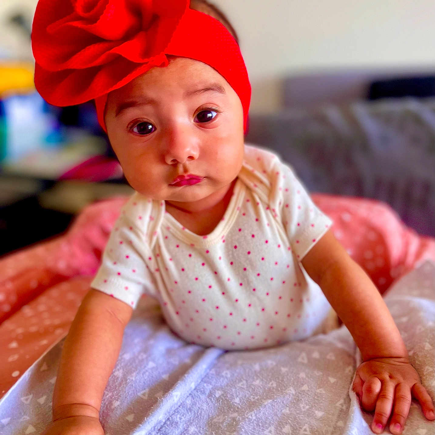 Gio is registered to the contest to win money with this photo: baby, blanket, child, closeup, couch, curious, cute, expression, face, flower, hands, indoors, infant, polka_dot_onesie, portrait, red_headband, skin, soft_toys, toddler, young_child