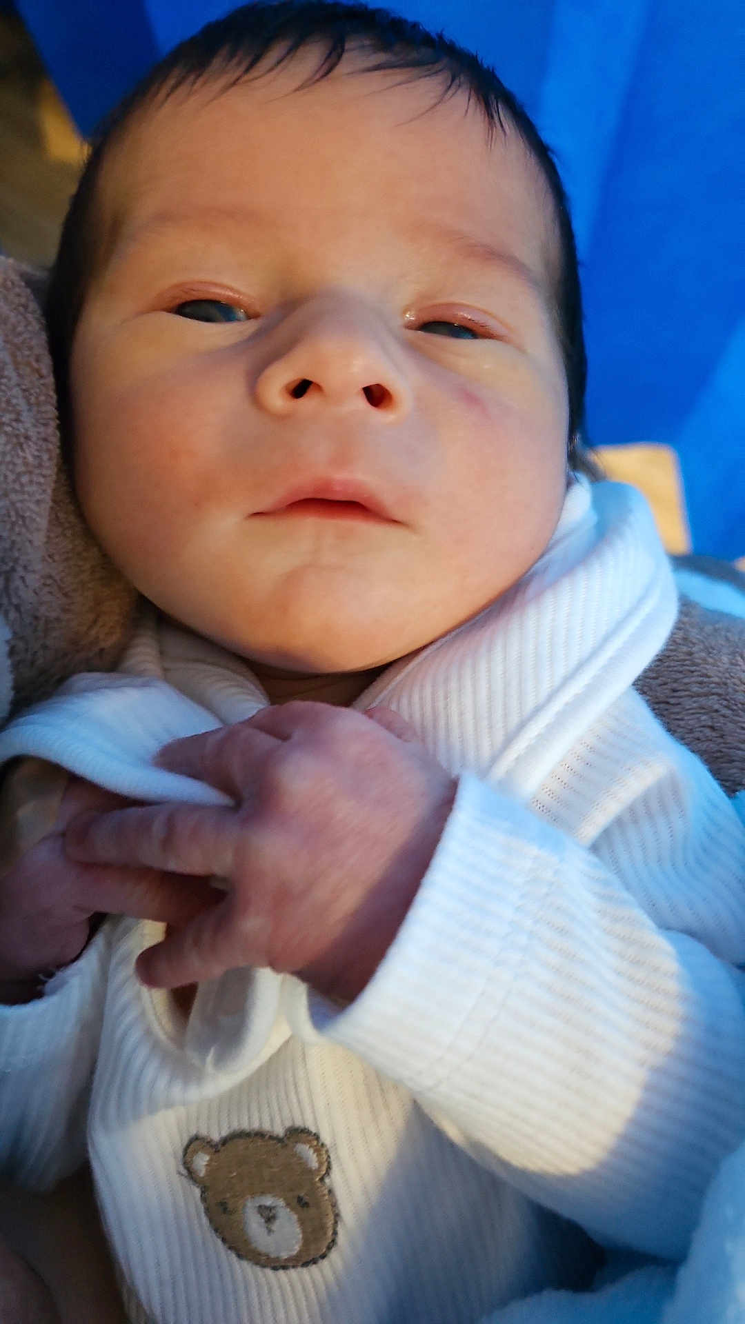 Anthony is registered to the contest to win money with this photo: baby, newborn, infant, close_up, face, hands, white_clothing, teddy_bear_patch, soft_texture, blue_background, cute, small_hands, portrait, indoors, child, baby_clothes, skin, person, newborn_baby, sleepy