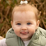 Cambrie is registered to the contest to win money with this photo: toddler, child, face, expression, tongue, bow, hair, vest, green, outdoor, nature, portrait, cute, baby, smile, warm_clothing, young_child, person, closeup, funny_face