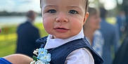 Finn is registered to the contest to win money with this photo: child, toddler, baby, face, person, clothing, vest, flower, boutonniere, hand, outdoor, blurred_background, nature, people, portrait, smile, short_hair, blue, white, holding