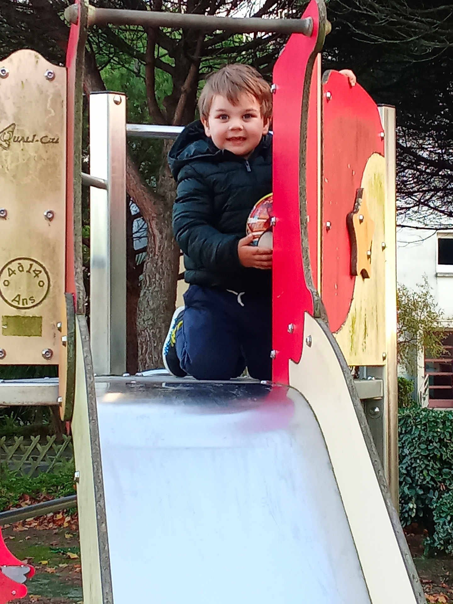 Milan participe au concours pour gagner de l'argent avec cette photo : child, playground, slide, ball, smiling, outdoor, tree, jacket, happy, play, park, boy, casual_clothing, daylight, recreation, fun, toy, nature, kneeling, sport_shoes