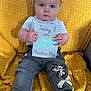 baby, blue_eyes, card, cartoon_pants, child, clothing, cute, expression, face, footwear_absent, grey_shirt, hands, indoor, legs, milestone, milestone_card, person, seated, texture, yellow_blanket