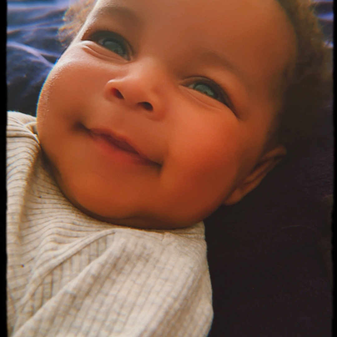 Kamari joined the competition — help win amazing prizes! baby, smiling, face, child, portrait, closeup, infant, cozy, striped_clothing, soft_texture, lying_down, happy, cute, blue_background, dark_surface, phone_screen, black_border, young_child, expression, warm_lighting