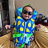 bed, blanket, casual, child, clothing, dog, furniture, hand, happy, indoor, life_jacket, open_door, person, pet, pineapple_pattern, playful, room, smile, sunglasses, toddler