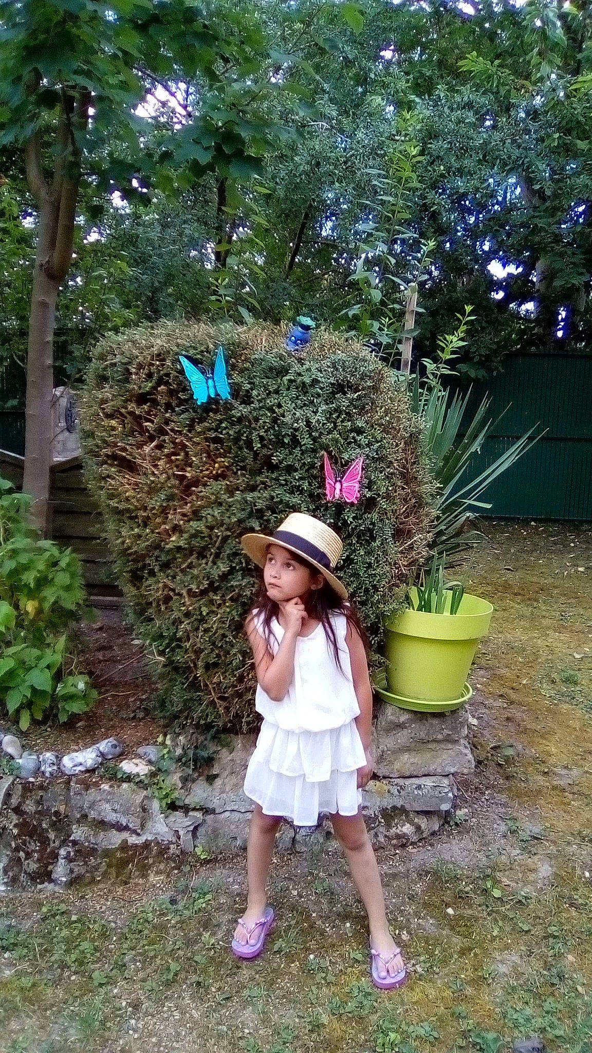 Tïana participe au concours pour gagner de l'argent avec cette photo : baby_toddler_clothing, backyard, clothing, costume_accessory, costume_hat, day_dress, dress, fedora, flowerpot, foot, garden, hat, headwear, home_fencing, person, sandal, shrub, sun_hat, yard