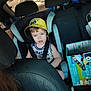 child, toddler, car_seat, seatbelt, yellow_hat, pokemon_hat, backseat, headrest, training_pants_box, cardboard_box, minions, vehicle_interior, portrait, facial_expression, young_child, car_window, clothing, seat, candid_shot, person
