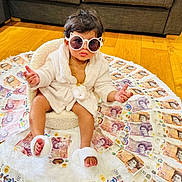 Trisha joined the competition — help win amazing prizes! baby, child, sunglasses, robe, money, british_pounds, floor, chair, wooden_floor, currency, indoor, plush, footwear, expression, sitting, cute, fashion, accessory, luxury, portrait