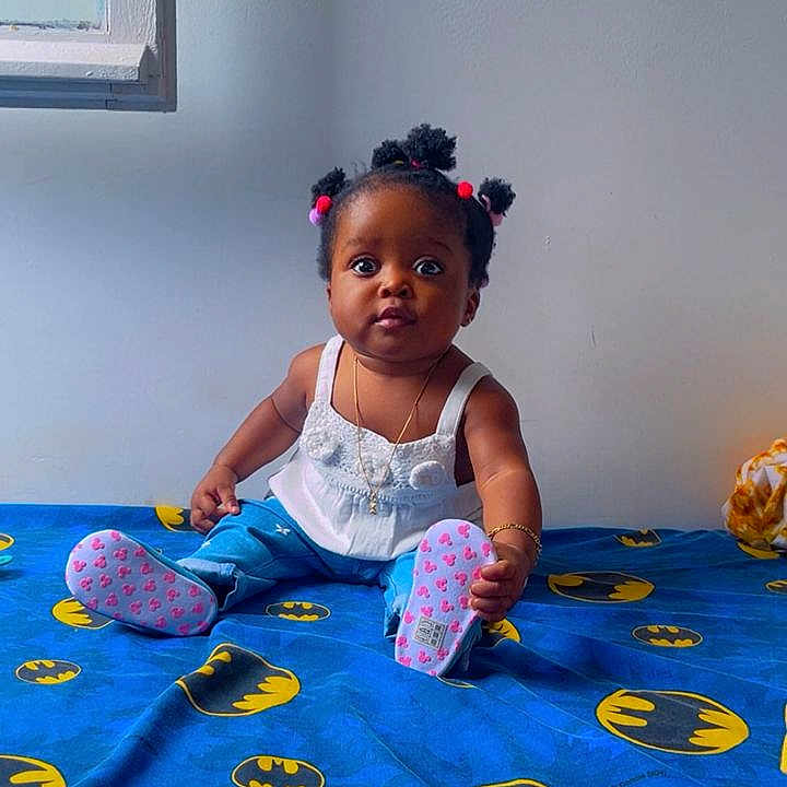 Aliah Saran participe au concours pour gagner de l'argent avec cette photo : baby, batman_logo, bed, blue_bedspread, blue_pants, bracelet, child, curious_expression, cute, floor, indoors, necklace, pigtails, pink_booties, portrait, sitting, wall, white_top, window, young_child