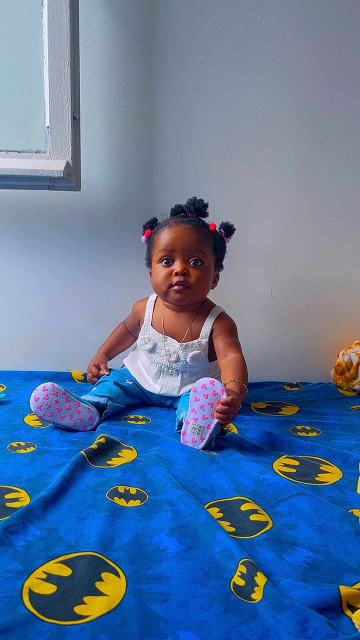 Aliah Saran participe au concours pour gagner de l'argent avec cette photo : baby, child, sitting, bed, batman_logo, blue_bedspread, pigtails, curious_expression, indoors, white_top, blue_pants, pink_booties, bracelet, necklace, window, wall, floor, cute, portrait, young_child