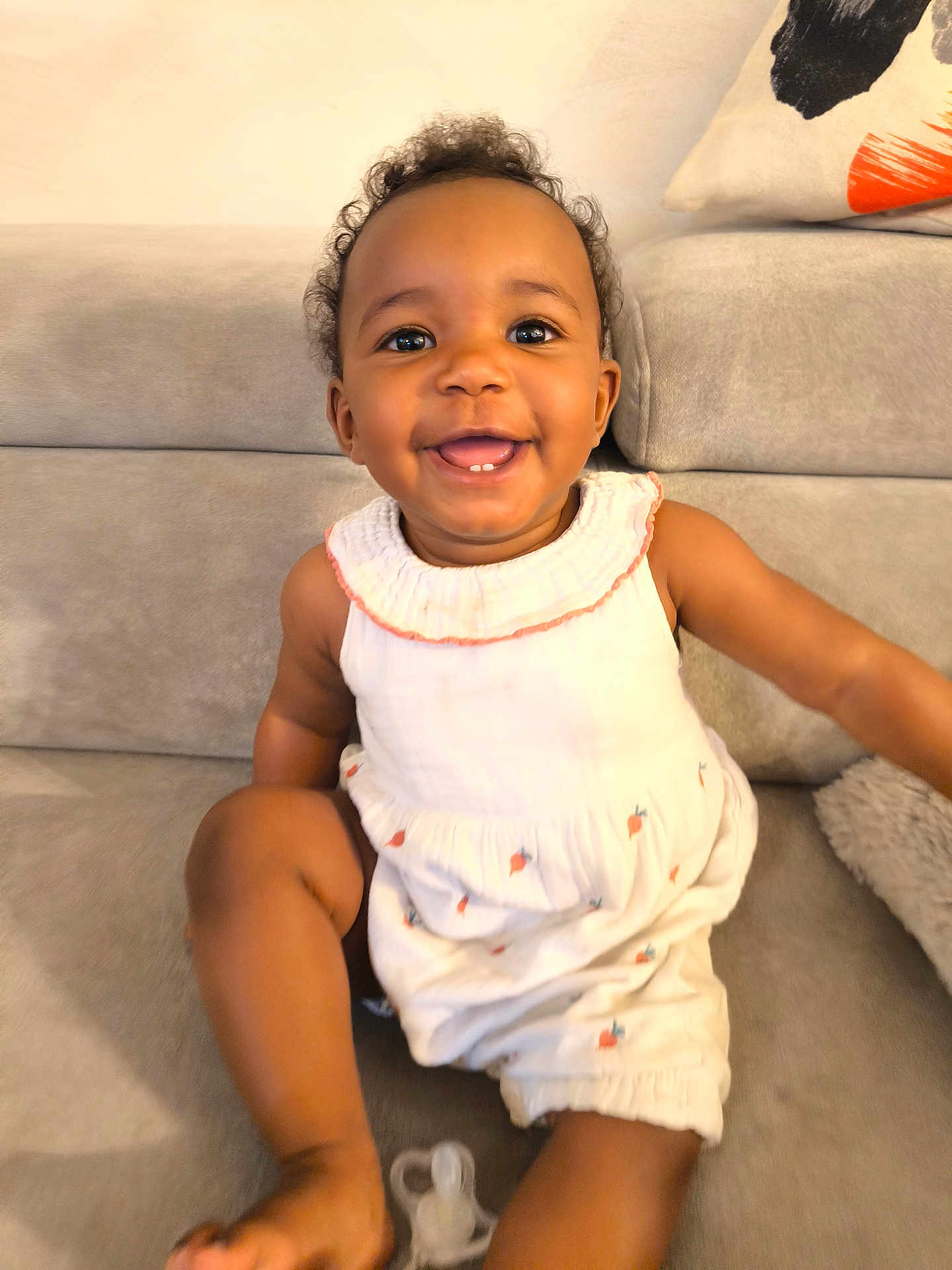 Mayvis a rejoint le concours — aidez-le/la à gagner de superbes lots ! baby, child, smile, face, sitting, couch, pacifier, white_outfit, curly_hair, happy, indoor, pillow, fabric, skin, infant, cute, portrait, one_person, young_child, home