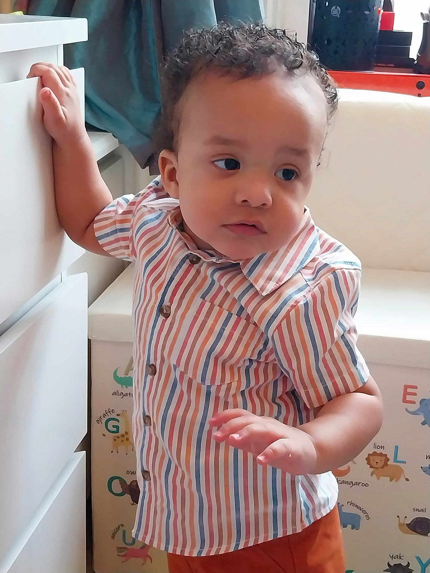 Aziah is registered to the contest to win money with this photo: toddler, child, striped_shirt, curly_hair, indoor, furniture, drawer, alphabet_wall, animal_illustrations, colorful, casual_clothing, young_child, standing, hand_on_drawer, looking_away, playful_background, cozy, portrait, home, cute