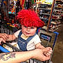 baby, child, shopping_cart, hat, red_feathers, denim_overall, white_shirt, tattoo, hand, store, shelves, toys, shopping, retail, face, blue_eyes, person, indoor, cute, infant
