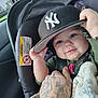 baby, infant, car_seat, hat, cap, new_york_yankees, hand, tattoo, blanket, smile, face, eyes, clothing, car_interior, seatbelt, child, person, holding, cute, portrait